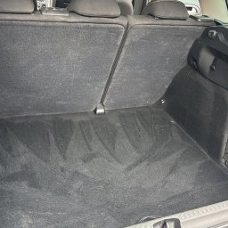 VILUX Jakub Gil - Wyprana tapicerka bagażnika samochodu, widoczne ślady po czyszczeniu na ciemnej wykładzinie. Siatka w bocznej części bagażnika.