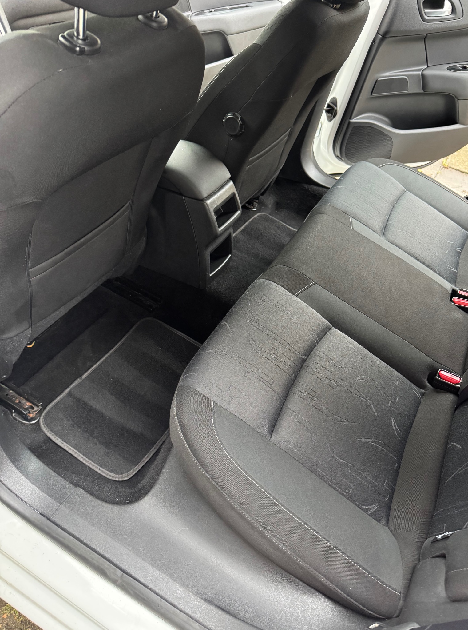 Wnętrze samochodu z czarną tapicerką, widoczne tylne siedzenia i dywaniki. Widoczne ślady użytkowania na tapicerce.