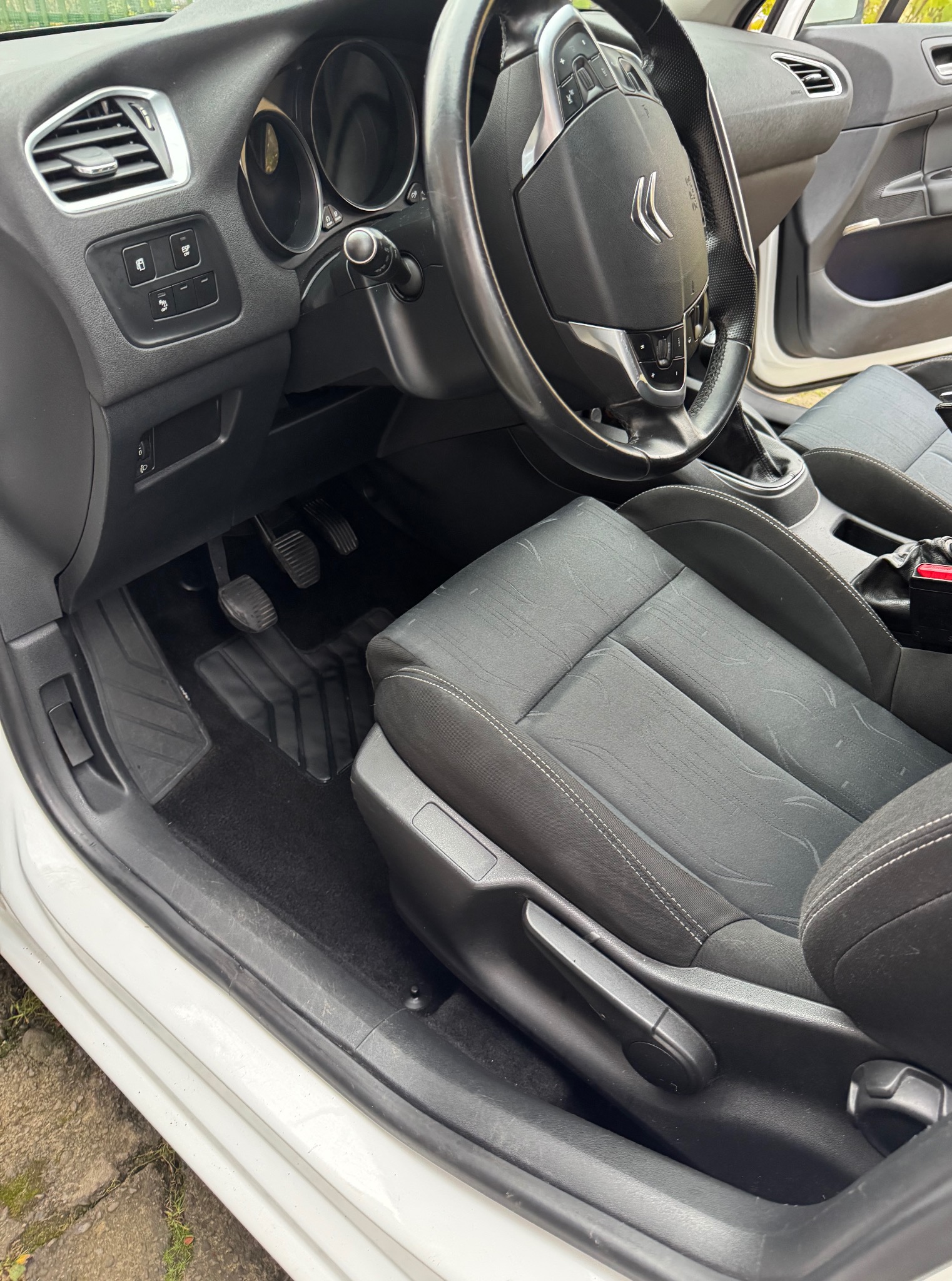 Wnętrze białego Citroena C4: czarna tapicerka fotela kierowcy, dywaniki, deska rozdzielcza i kierownica z logo. Widoczne pedały i dźwignia regulacji fotela.