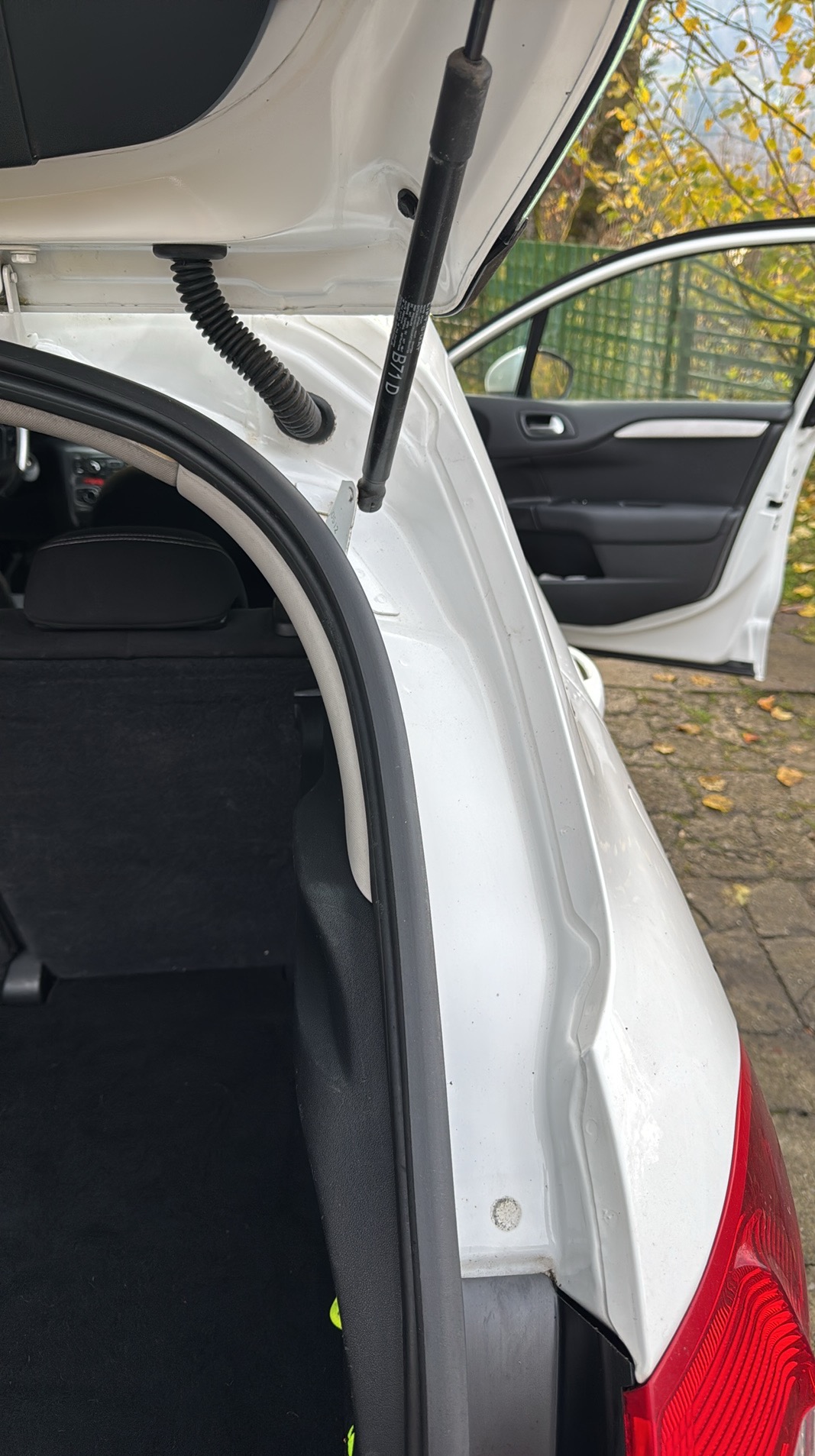 Otwarty bagażnik białego samochodu z widocznym wnętrzem i elementami karoserii, drzwi pojazdu otwarte na tle jesiennych drzew.