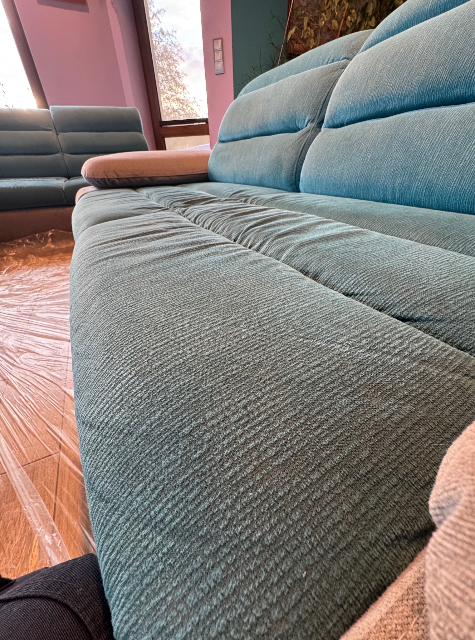 Turkusowa kanapa z poduszką, zabezpieczona folią na podłodze. Widok z boku, ukazujący detale faktury tkaniny i staranne ułożenie poduszki.