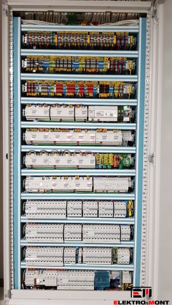 Rozdzielnica elektryczna w szafie, z precyzyjnie ułożonymi kablami i modułami, widok z przodu, oświetlenie górne, etykiety na elementach.