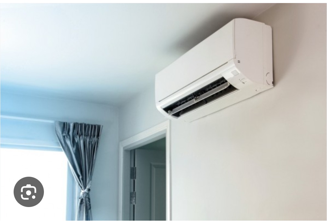 Biała klimatyzacja ścienna w jasnym pokoju z zasłoniętym oknem i drzwiami w tle. Widok z dołu, neutralne oświetlenie.