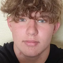 Portret młodego mężczyzny z kręconymi włosami i jasnymi oczami, ubrany w czarną koszulkę. Widoczny fragment śladu na szyi. Neutralne tło.