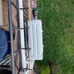 MAGELECTRIC - Zamontowana jednostka zewnętrzna klimatyzacji na balkonie z widokiem na zieleń. Widoczne elementy konstrukcyjne balkonu i fragment elewacji budynku. Ujęcie z góry.