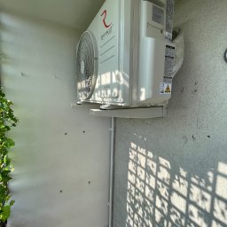 MAGELECTRIC - Biały klimatyzator Rotenso HIRO zamontowany na ścianie balkonu, widoczne rury instalacyjne i cień kraty. Świeża zieleń roślin w lewym dolnym rogu kadru.