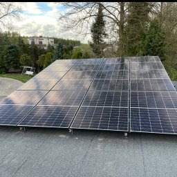 MAGELECTRIC - Panele fotowoltaiczne na dachu budynku, odbijające niebo i chmury. Widoczne mocowania i otoczenie z zielenią. Ujęcie z góry, lekko z boku.