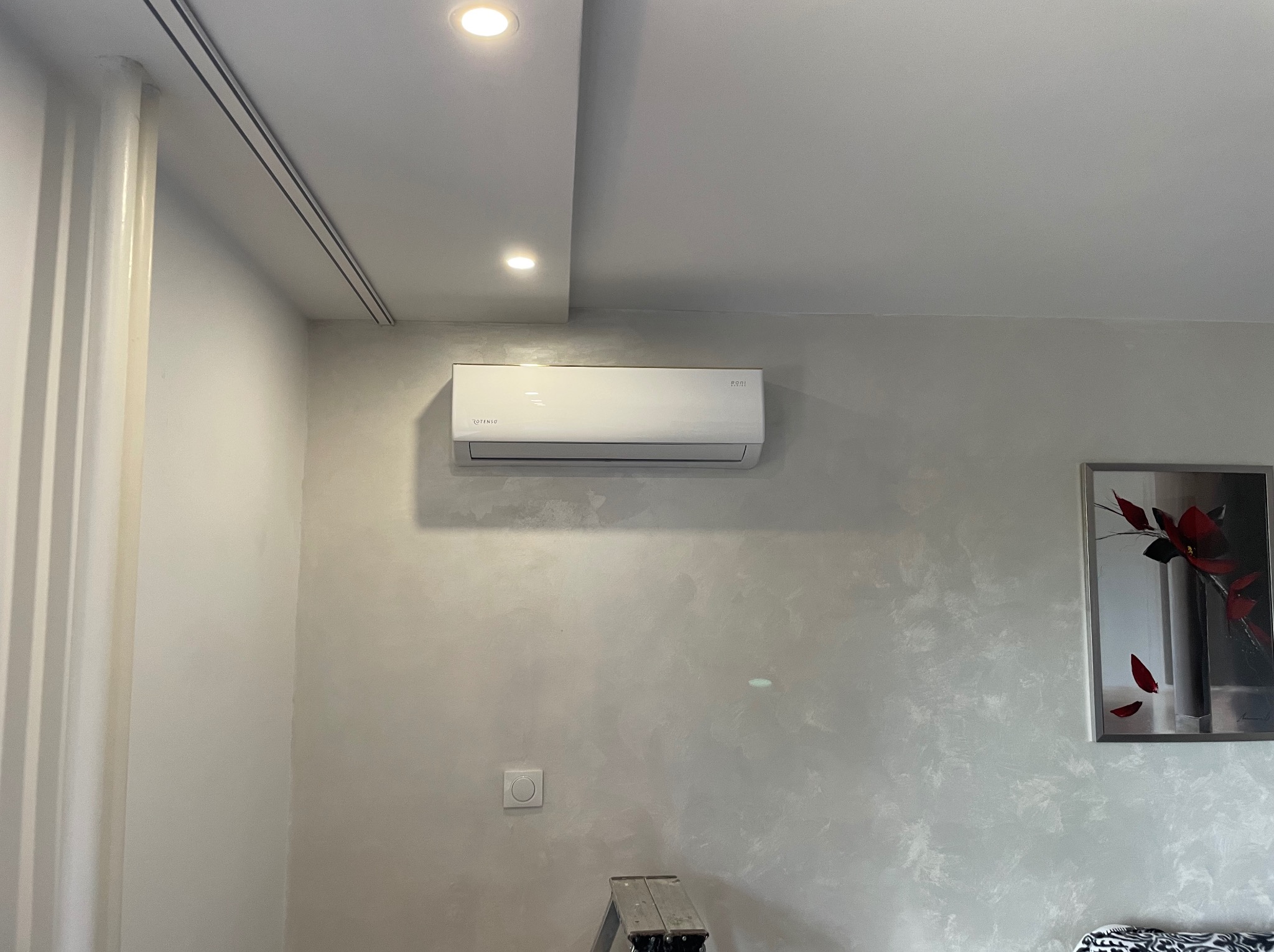 Biały klimatyzator Rotenso zamontowany na ścianie o dekoracyjnej fakturze, widoczny fragment drabiny i obraz w ramie. Oświetlenie sufitowe punktowe.