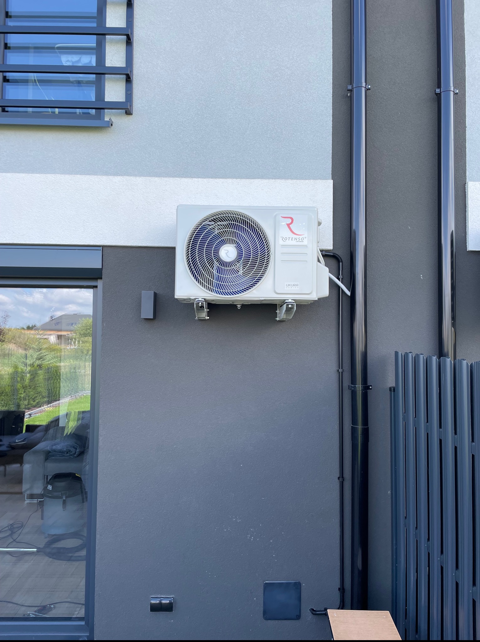 Biała jednostka zewnętrzna klimatyzacji Rotenso zamontowana na szarej ścianie budynku, widoczne rury odprowadzające skropliny i fragment okna z odbiciem wnętrza.