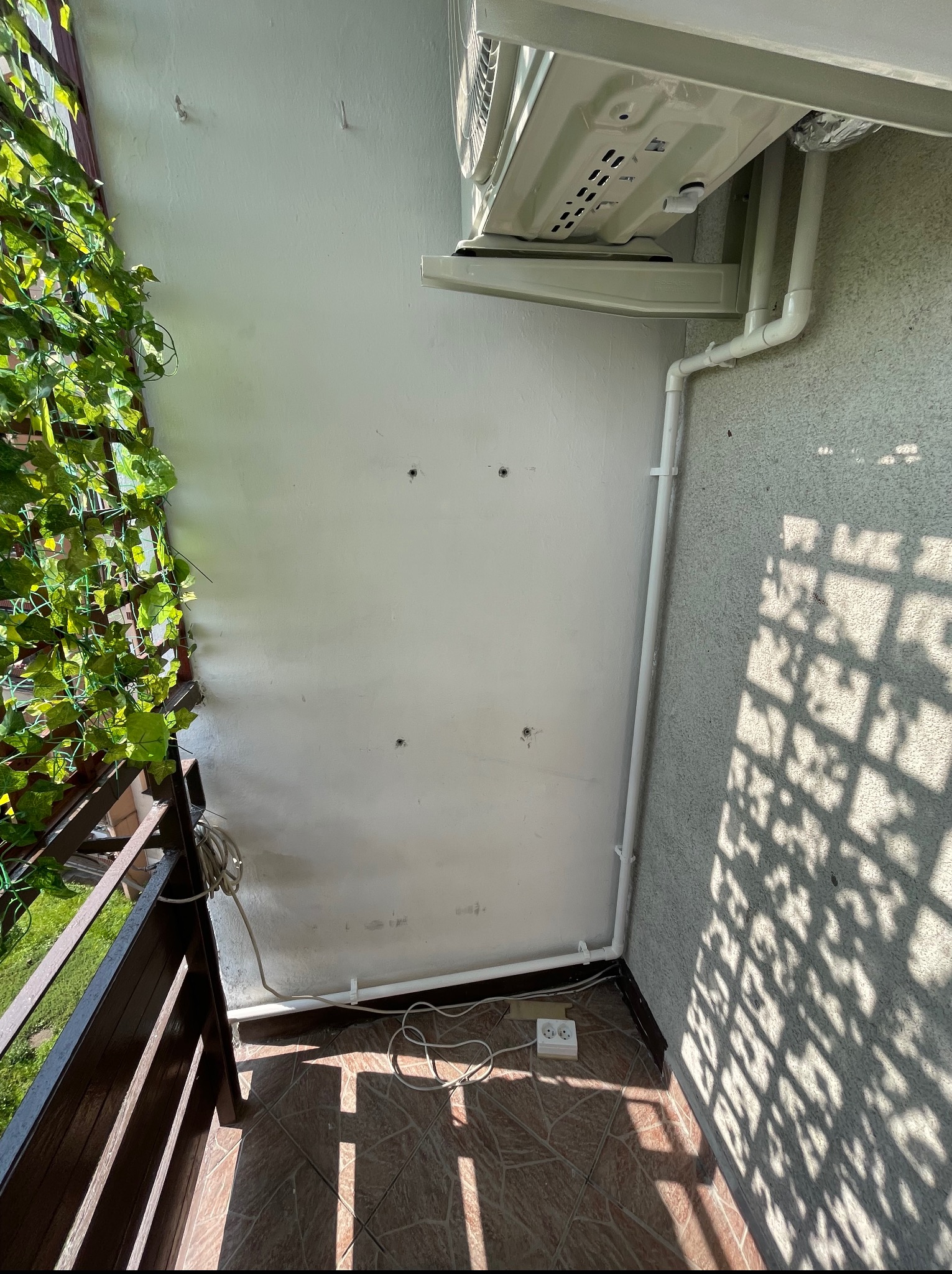 Biała jednostka klimatyzacji zamontowana na balkonie, widoczne rury odprowadzające. Na ścianie ślady po wierceniu, balkon z roślinami i gniazdko elektryczne.