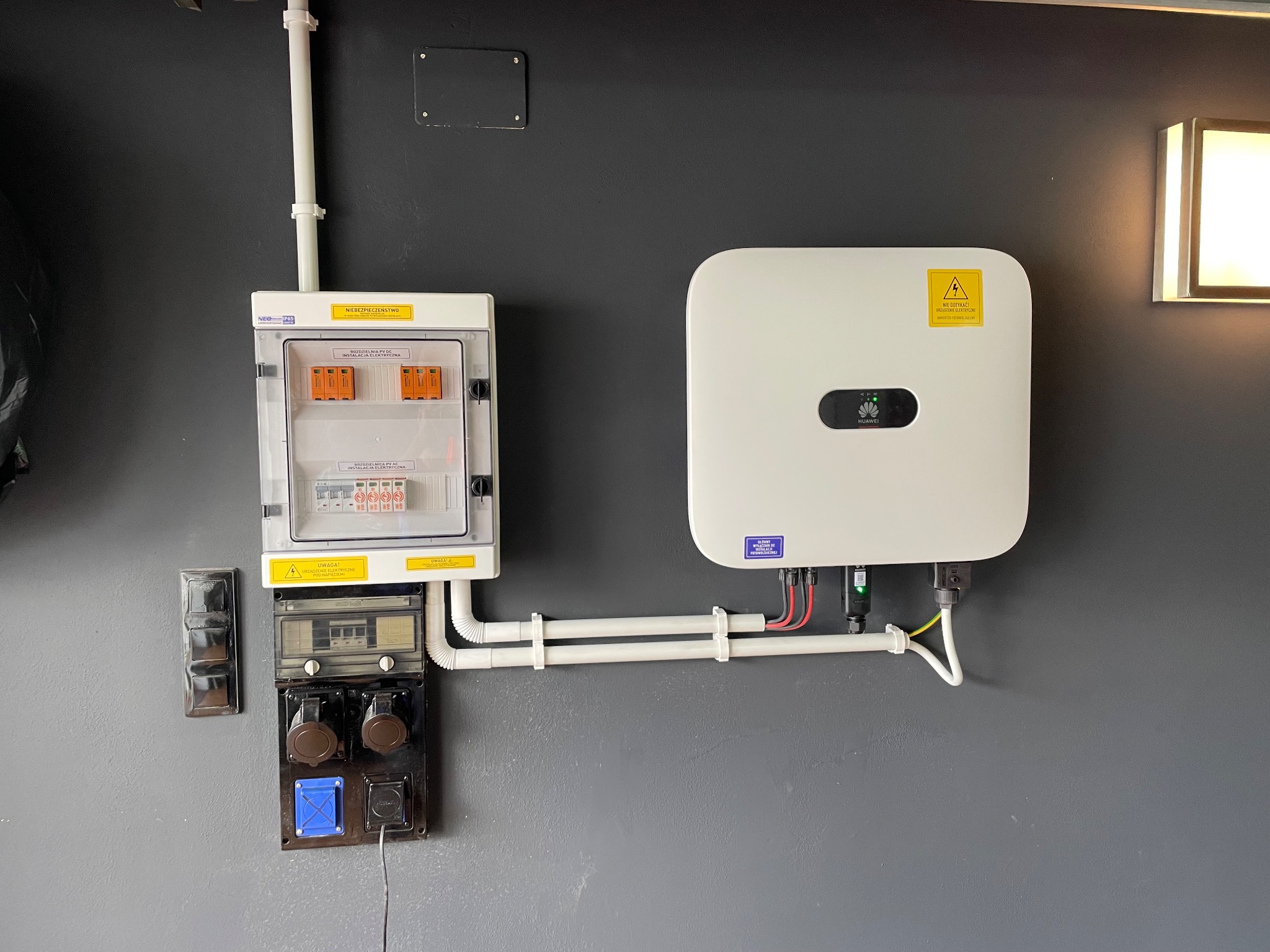 Biały inwerter Huawei i rozdzielnica PV AC na szarej ścianie. Widoczne przewody i zabezpieczenia. Element instalacji fotowoltaicznej wewnątrz budynku.