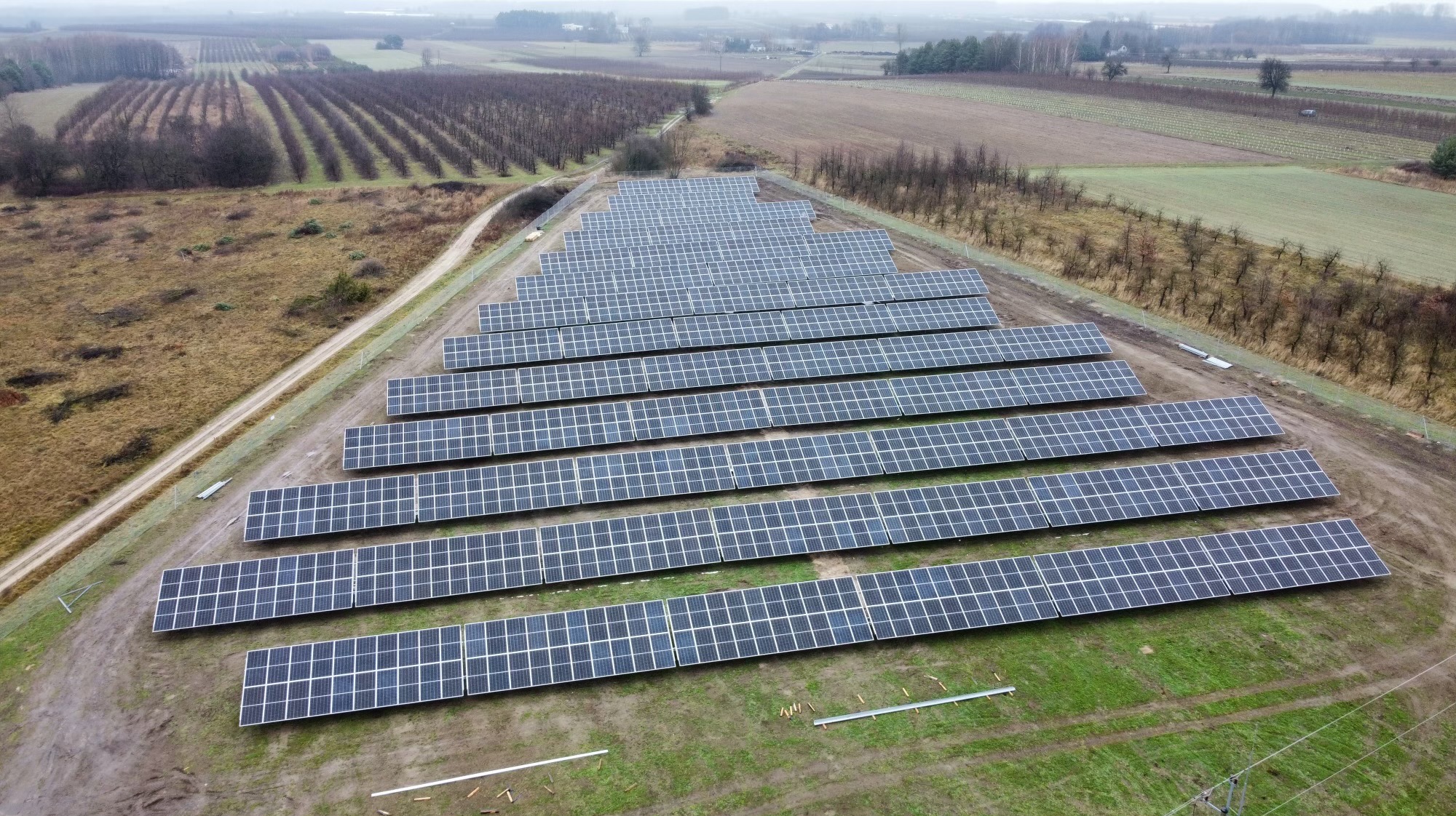 Rozległa farma fotowoltaiczna z panelami ułożonymi rzędami na polu, widok z góry, w tle drzewa i pola uprawne w pochmurny dzień. Widoczne elementy konstrukcyjne.