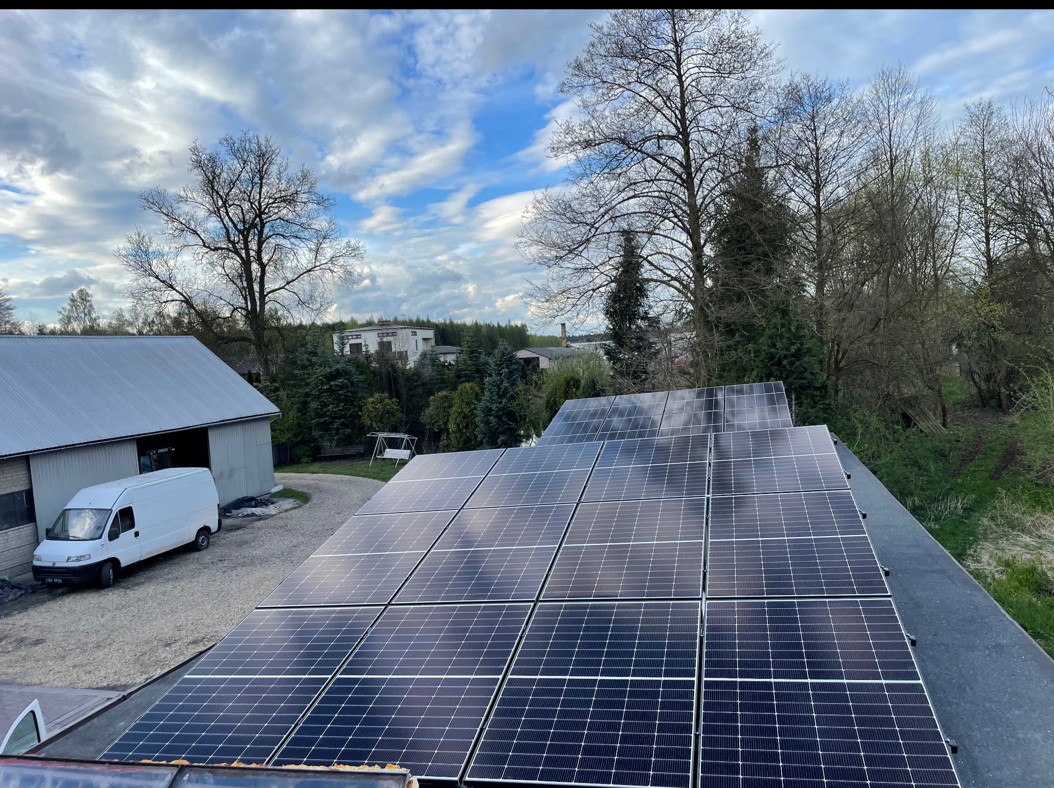 Panele fotowoltaiczne na dachu budynku w Krakowie, odbijające błękitne niebo z chmurami. W tle budynki i drzewa, a obok biały van. Widok z góry.