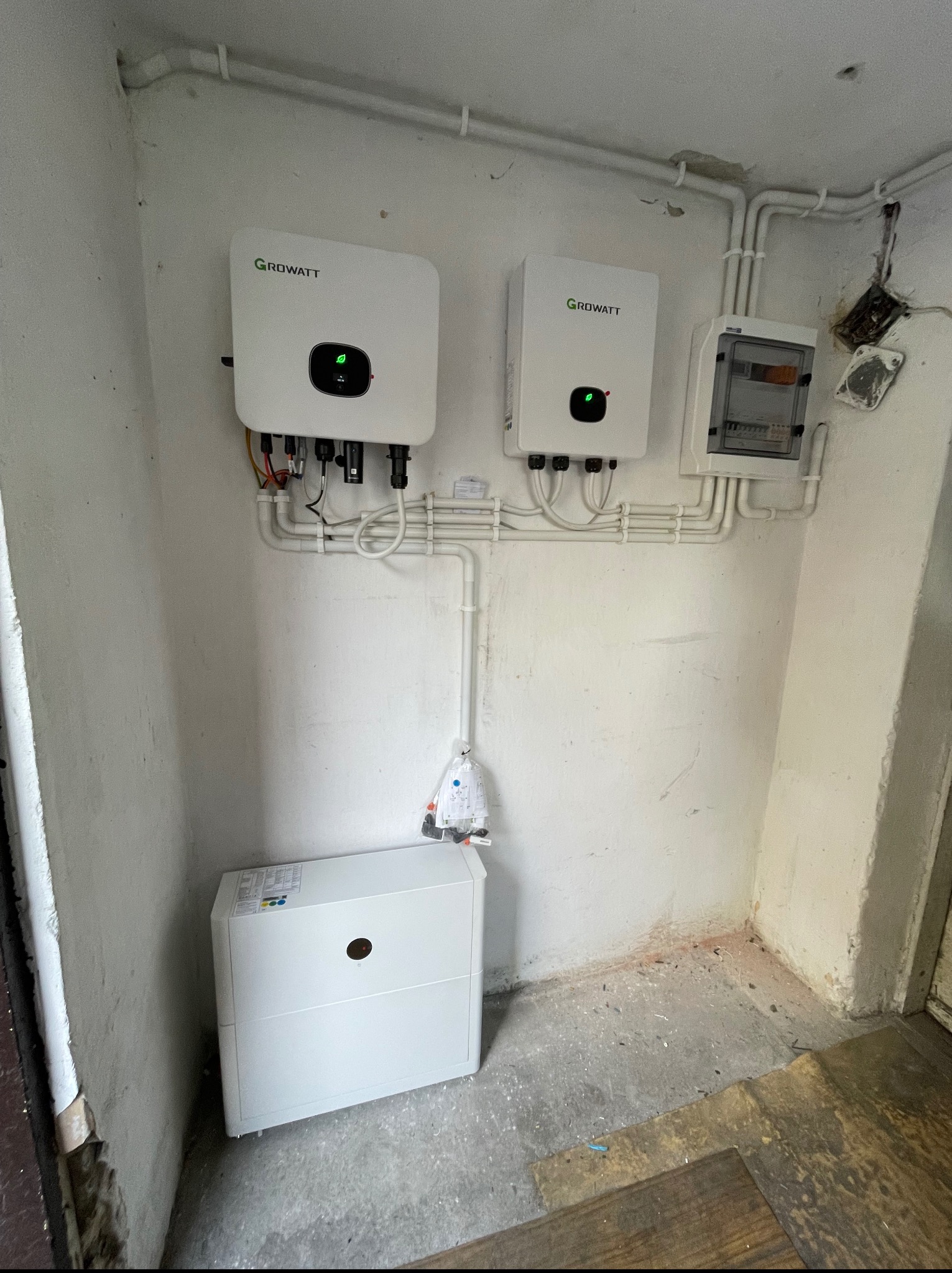 Dwa inwertery Growatt i skrzynka elektryczna zamontowane na białej ścianie w pomieszczeniu. Widoczne białe rurki instalacyjne i biała obudowa akumulatora na podłodze.