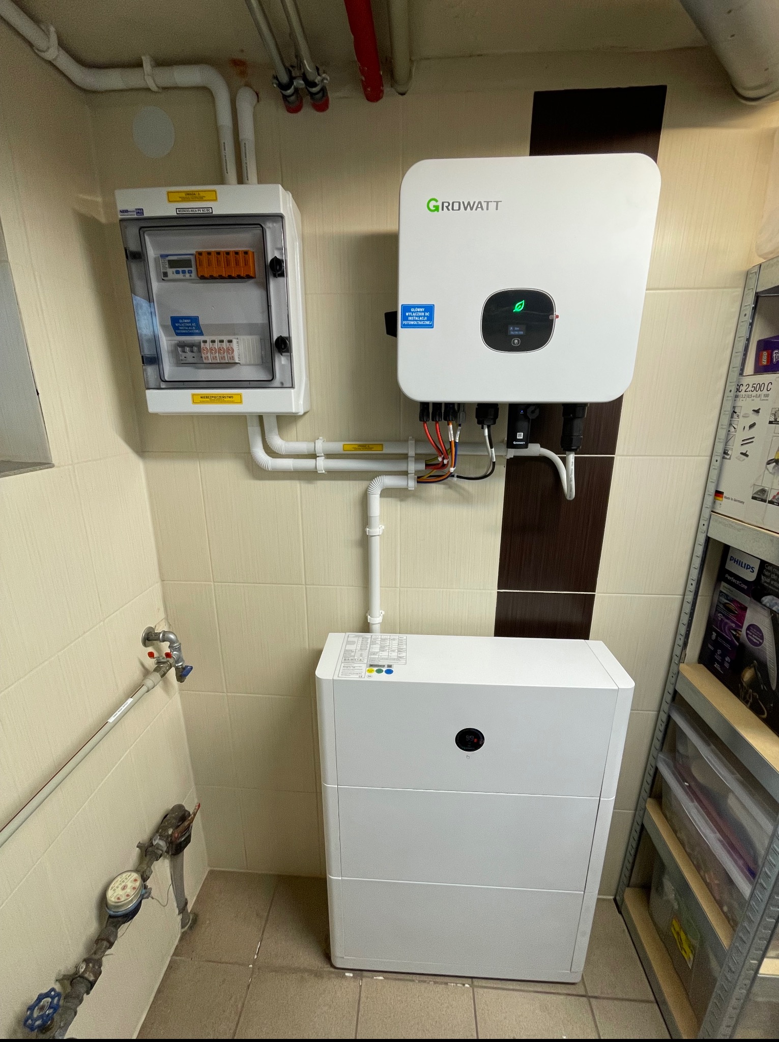 Zainstalowany system fotowoltaiczny w pomieszczeniu: biały inwerter Growatt, rozdzielnica elektryczna i akumulator na tle beżowych płytek. Widoczne rury i licznik wody.