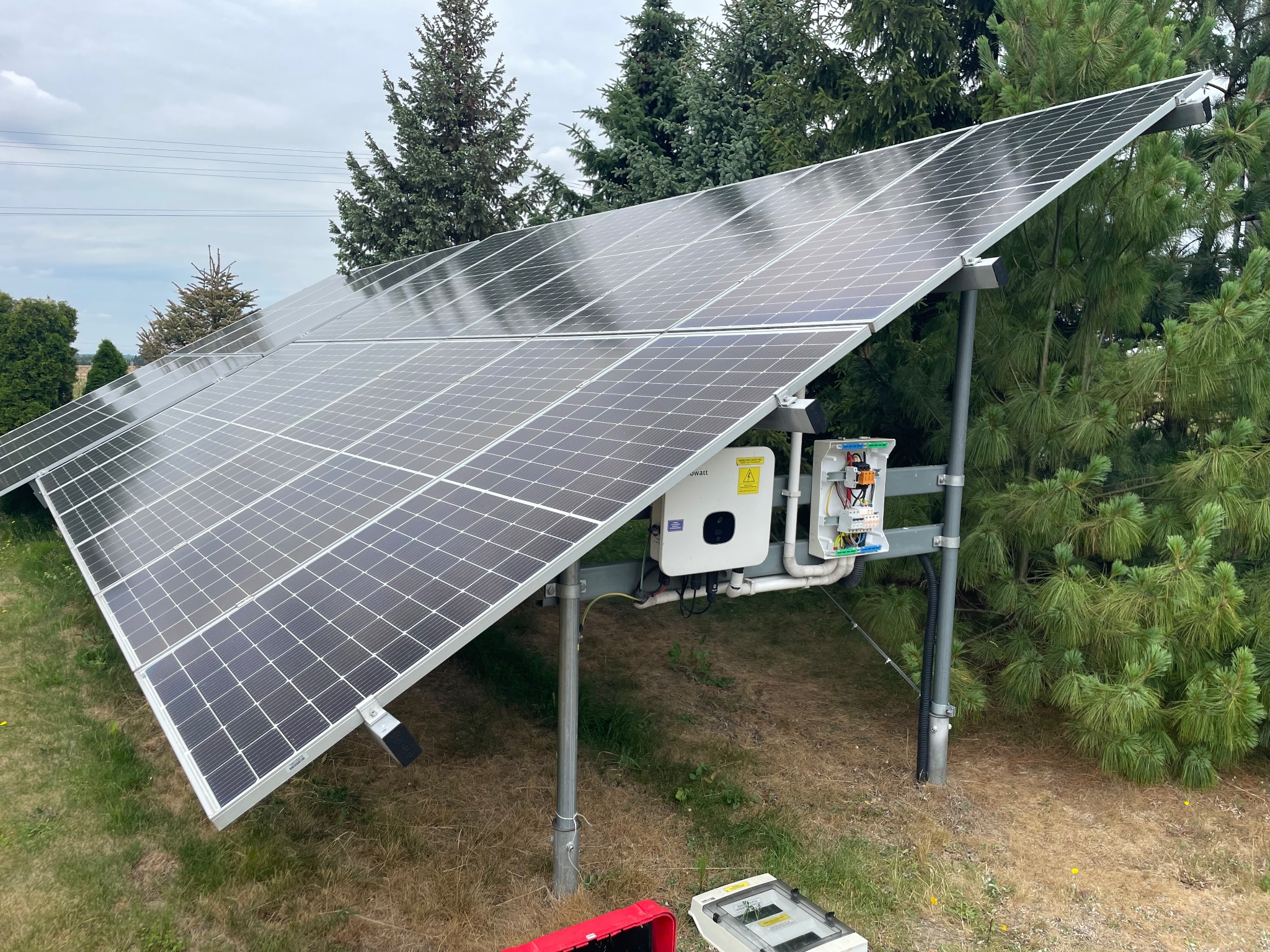Panel fotowoltaiczny na tle zieleni, z widocznym inwerterem i skrzynką elektryczną. Instalacja na trawniku, częściowo widoczne narzędzia.