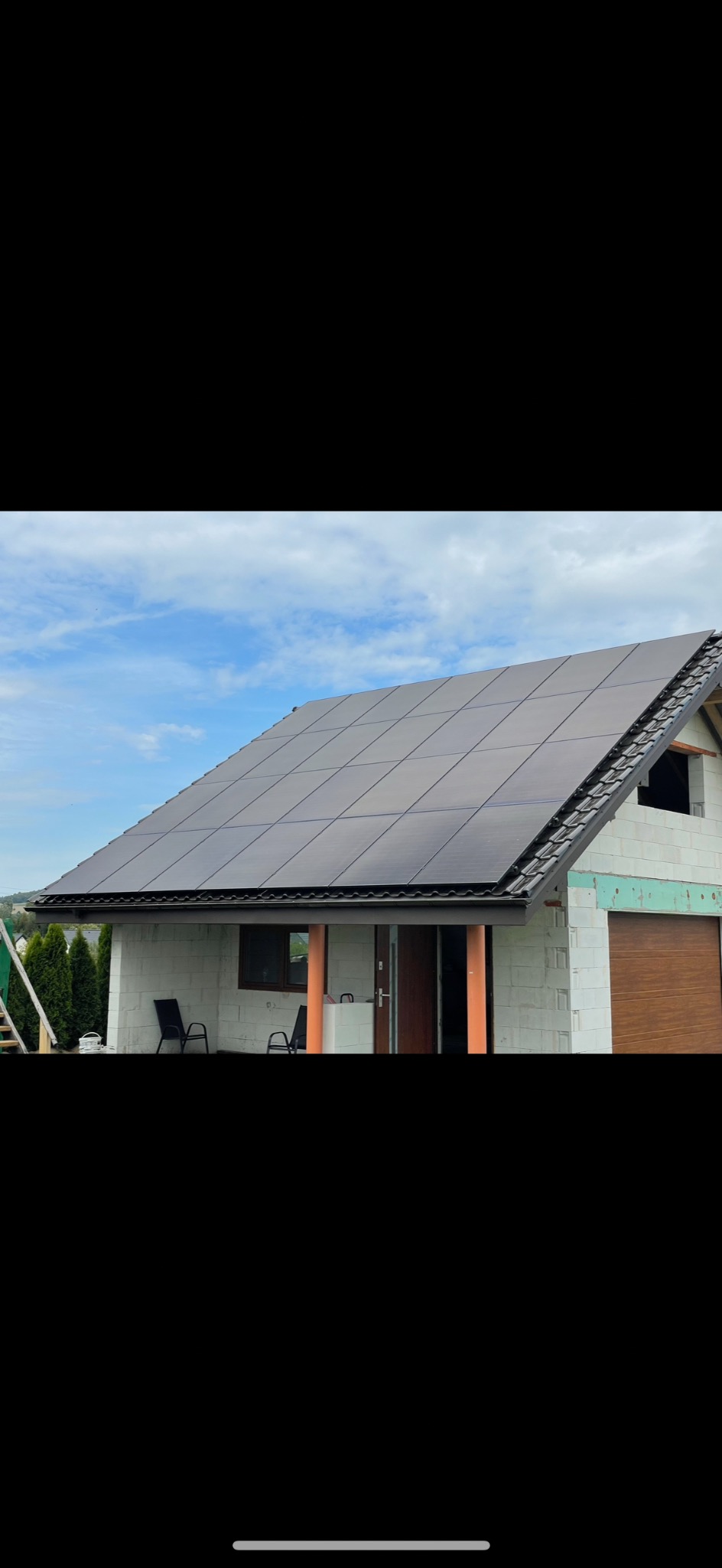 Dom w trakcie budowy z zamontowanymi panelami fotowoltaicznymi na dachu. Widoczne białe ściany z pustaków i brązowa brama garażowa.