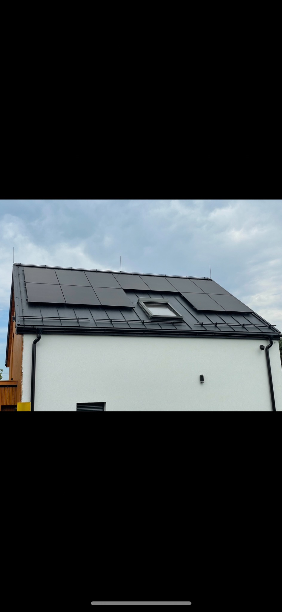 Biały dom z czarnym dachem i panelami fotowoltaicznymi. Widoczne okno dachowe i piorunochrony. Pochmurne niebo w tle.