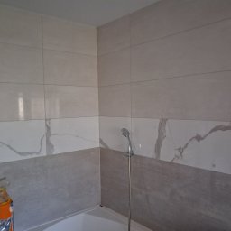 Waldemar Tomczewski - Wykończona łazienka: ściany wyłożone płytkami w dwóch odcieniach szarości, z motywem marmuru na górnej części. Widoczny prysznic i fragment wanny.