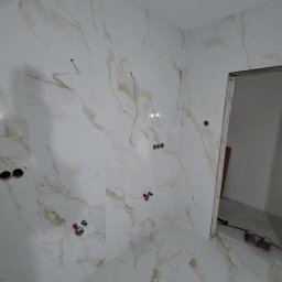 Waldemar Tomczewski - Wykończona łazienka z białymi płytkami imitującymi marmur, widoczne wystające kable i rury, przygotowane do montażu armatury. Otwór drzwiowy bez ościeżnicy.