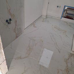 Waldemar Tomczewski - Białe, marmurkowe płytki podłogowe i ścienne w łazience, widoczne zawory i rozdzielacz instalacji. Ukończone prace, jasne wnętrze.