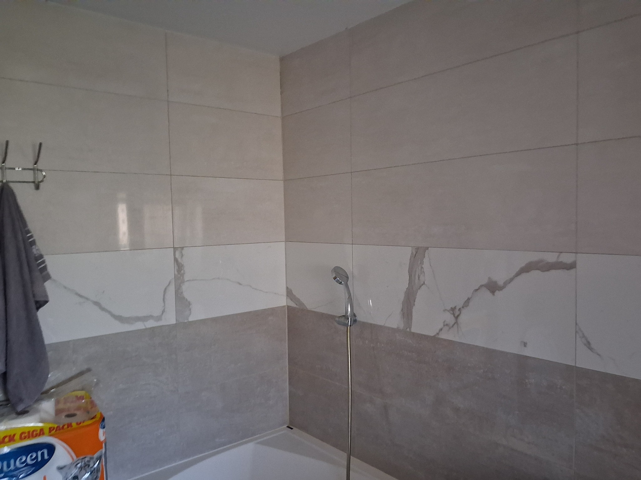 Wykończona łazienka: ściany wyłożone płytkami w dwóch odcieniach szarości, z motywem marmuru na górnej części. Widoczny prysznic i fragment wanny.