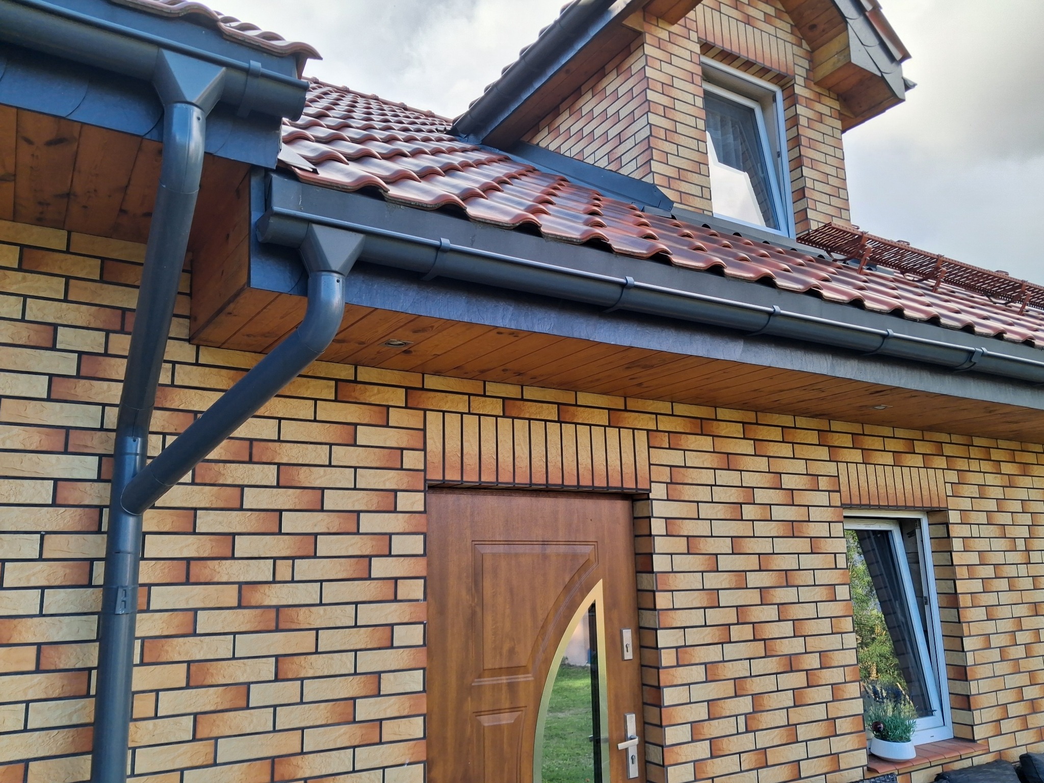 Fragment elewacji domu z klinkieru: brązowe cegły, ciemne rynny, drewniana podbitka dachu, okno z parapetem i brązowe drzwi wejściowe. Widok z dołu.