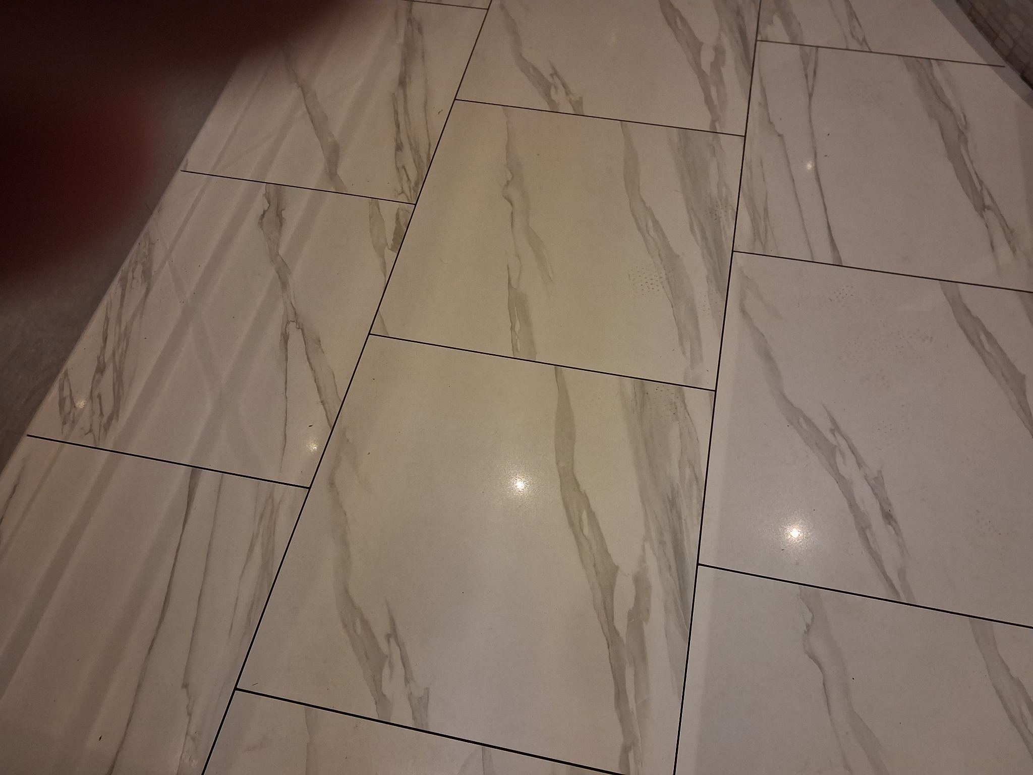 Ułożone płytki podłogowe imitujące marmur, z widocznymi fugami. Jasne, beżowo-szare odcienie z charakterystycznym wzorem żyłkowania. Ujęcie pod kątem.