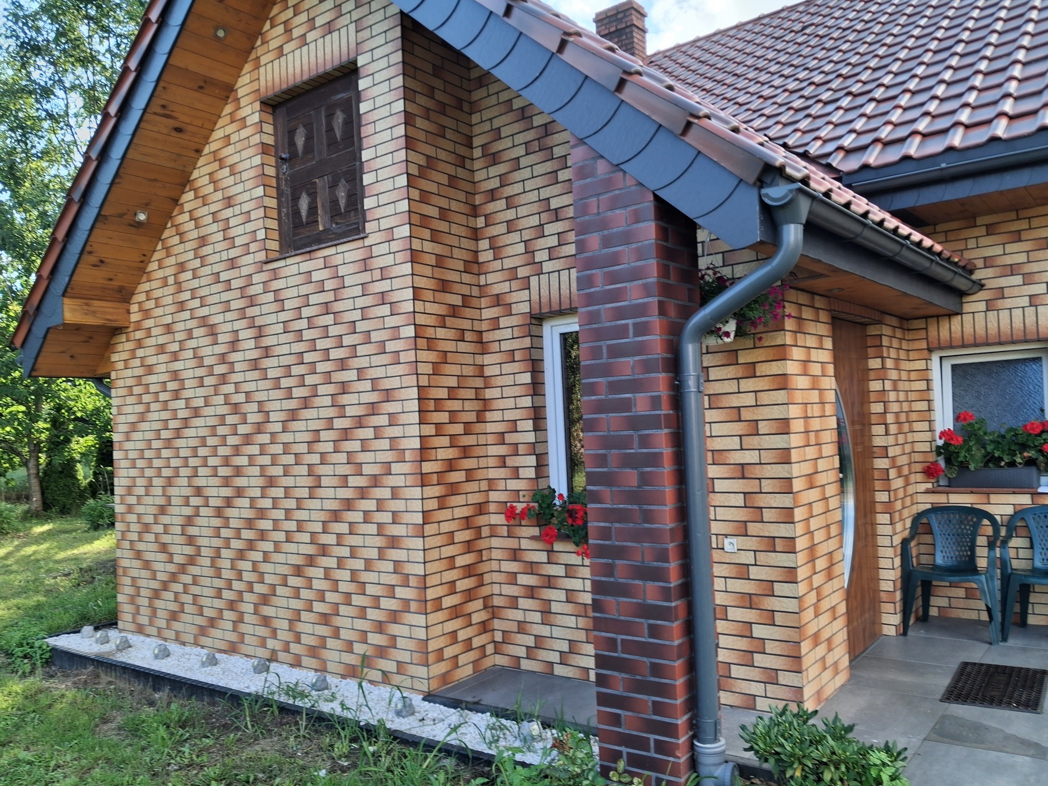 Dwukolorowa elewacja domu z cegły klinkierowej, z widocznymi oknami z kwiatami, rynną i fragmentem dachu. Ujęcie z poziomu gruntu, podkreślające detale wykończenia.
