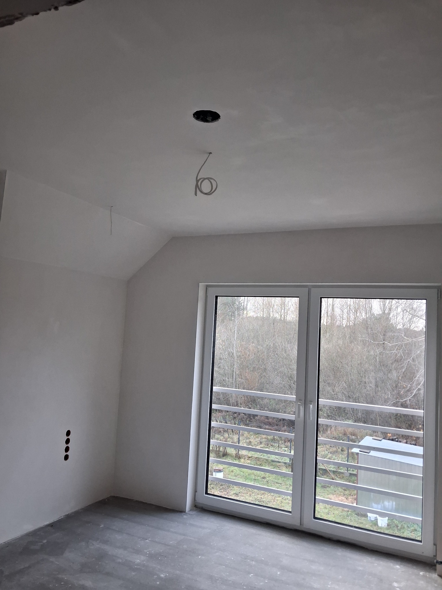 Pokój po szpachlowaniu, ściany i sufit na biało, widoczne otwory na gniazdka elektryczne i wyprowadzone kable oświetleniowe. Okno balkonowe z widokiem na zewnątrz.