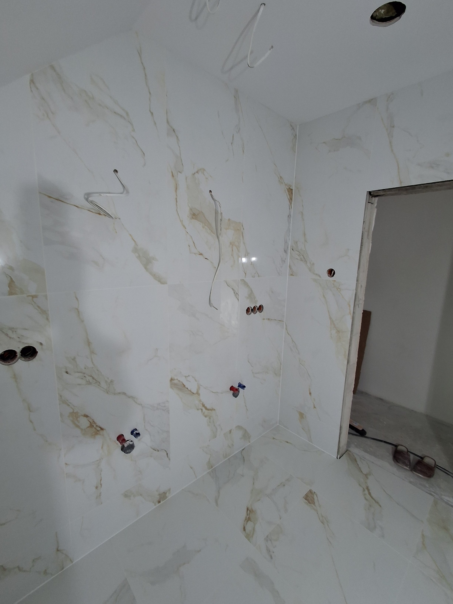 Wykończona łazienka z białymi płytkami imitującymi marmur, widoczne wystające kable i rury, przygotowane do montażu armatury. Otwór drzwiowy bez ościeżnicy.