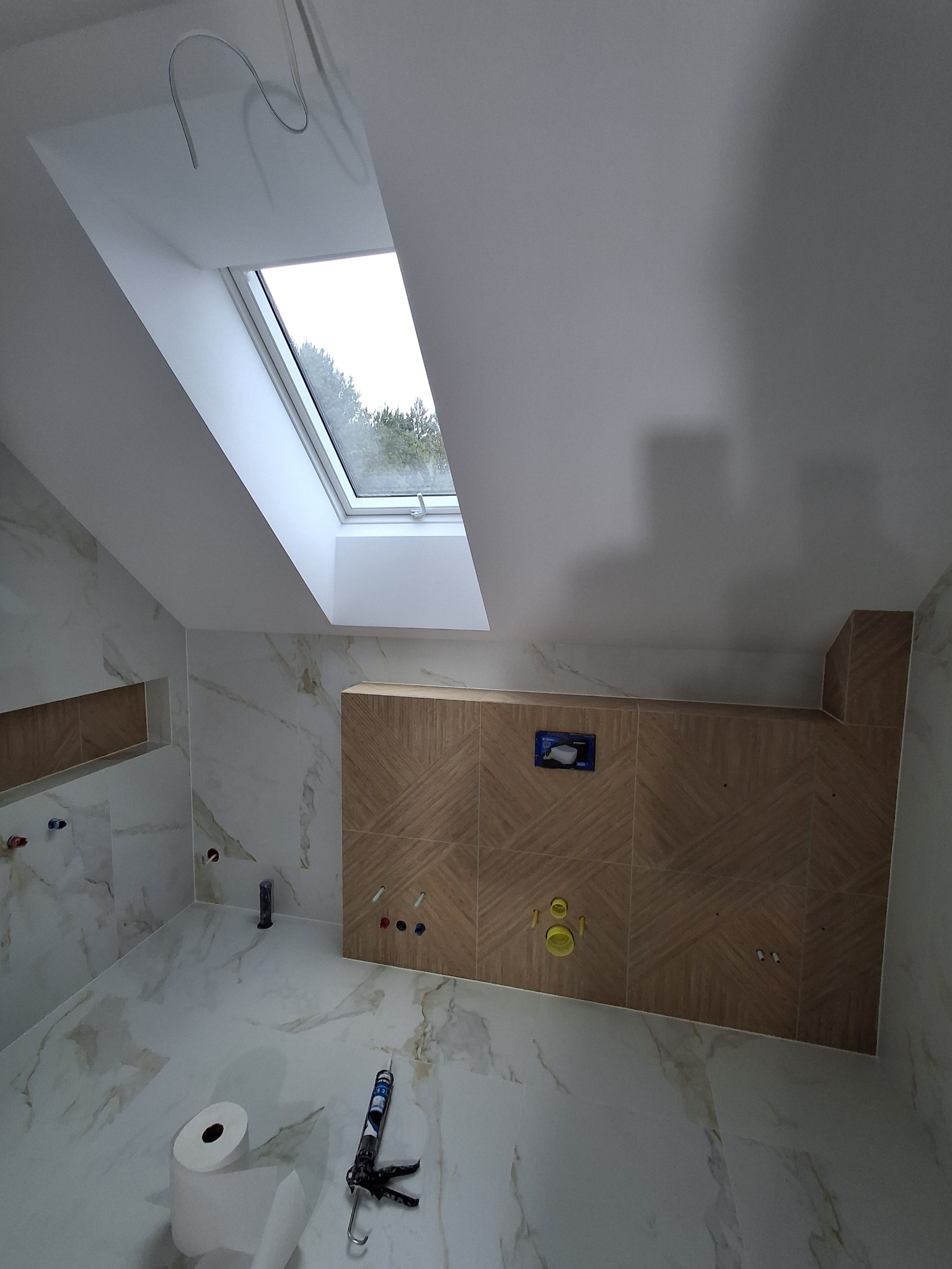 Wykończona łazienka na poddaszu z oknem dachowym. Marmurowe płytki na ścianach i podłodze, zabudowa z płytek imitujących drewno. Widoczne narzędzia glazurnicze.