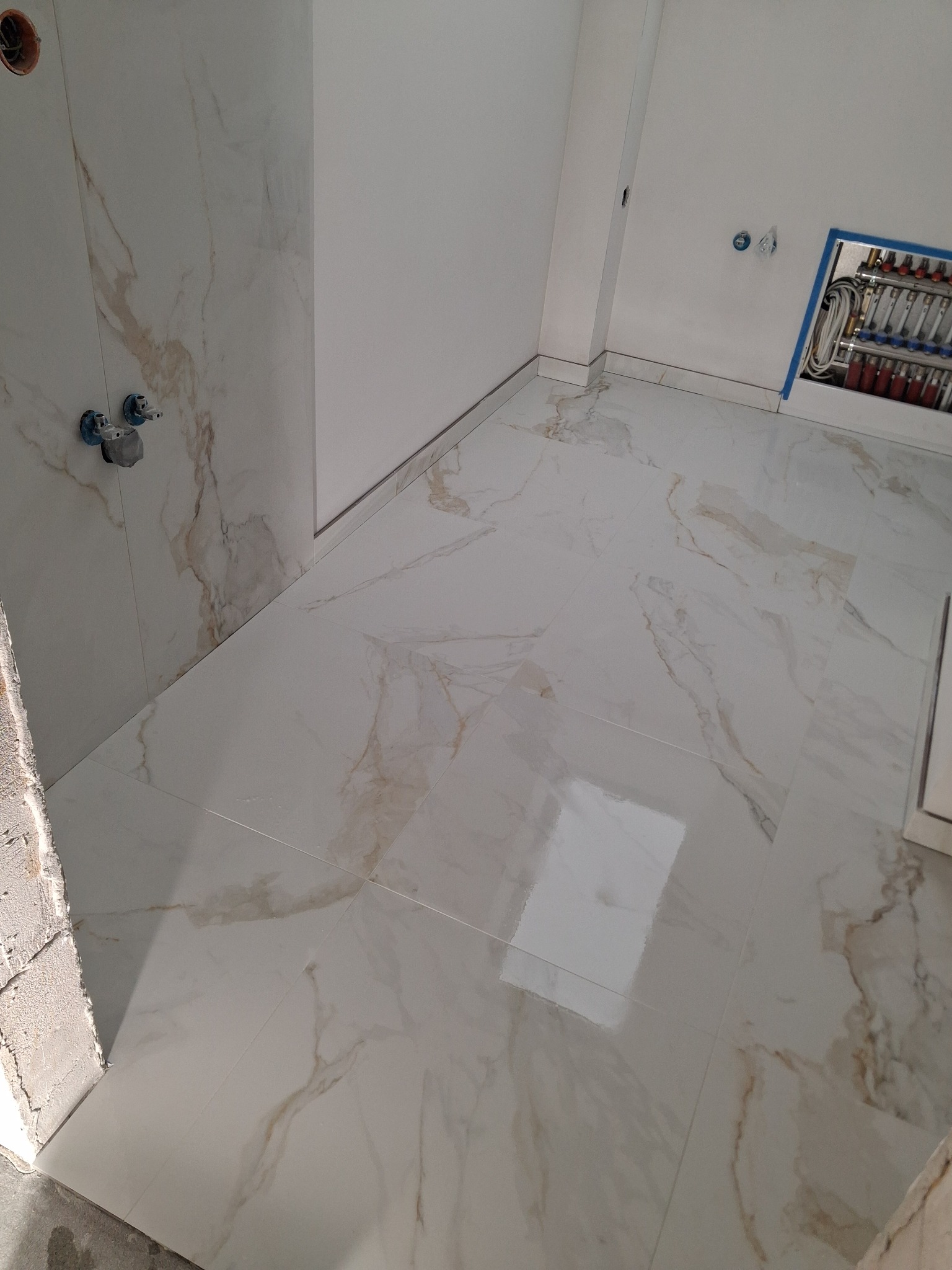 Białe, marmurkowe płytki podłogowe i ścienne w łazience, widoczne zawory i rozdzielacz instalacji. Ukończone prace, jasne wnętrze.