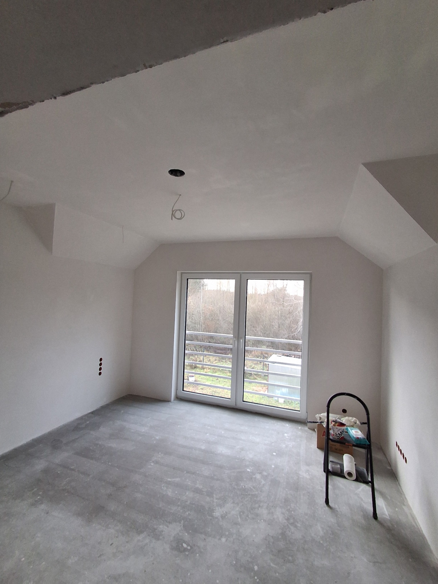 Wnętrze poddasza po gładziowaniu ścian i sufitu w Barczewie. Widoczne okno balkonowe, przygotowane otwory na gniazdka i drabina malarska.