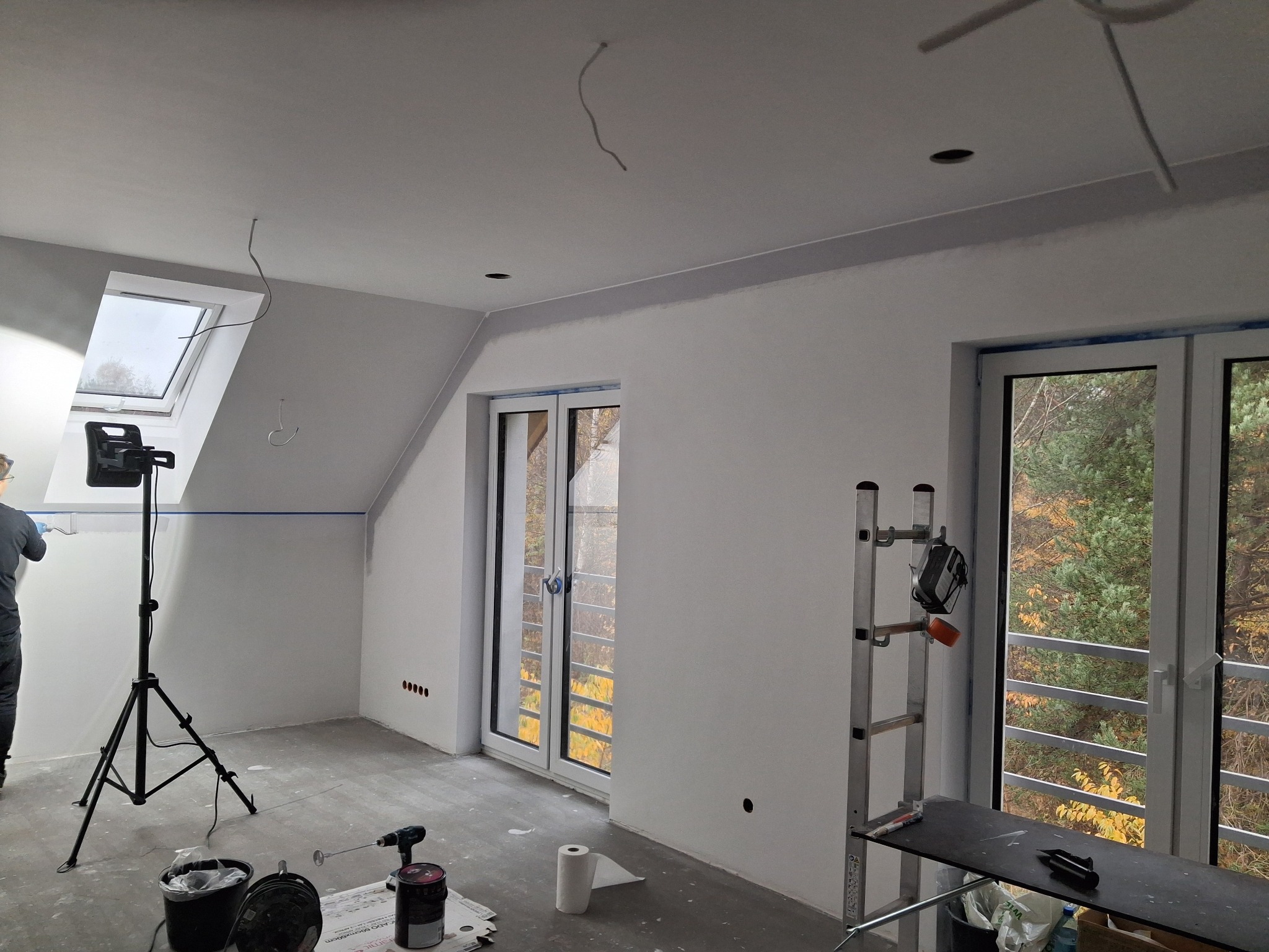Wnętrze w trakcie malowania: ściany na biało, widoczne okno dachowe, drabina, lampa na statywie i narzędzia malarskie. Świeżo pomalowane ściany w pokoju na poddaszu.