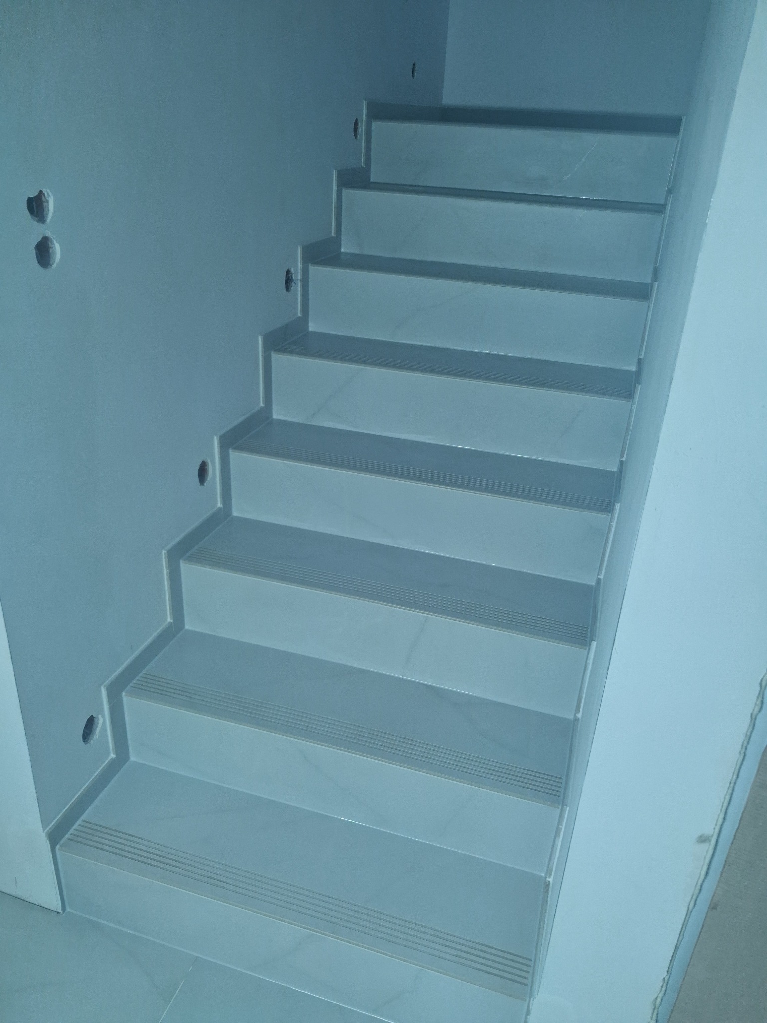 Wykończone schody wewnątrz budynku, wyłożone jasnymi, błyszczącymi płytkami z antypoślizgowymi rowkami. Widoczne otwory na oświetlenie w ścianie.