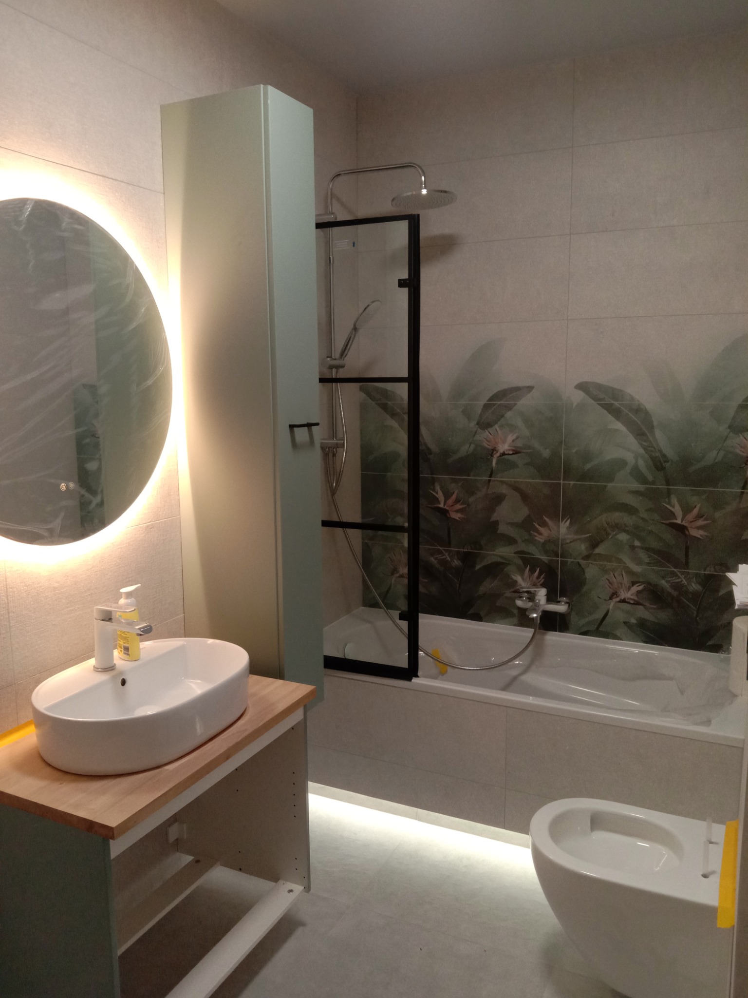 Jasna łazienka z owalną umywalką na drewnianym blacie, podświetlanym lustrem LED, kabiną prysznicową z czarną ramą i płytkami z motywem roślinnym.