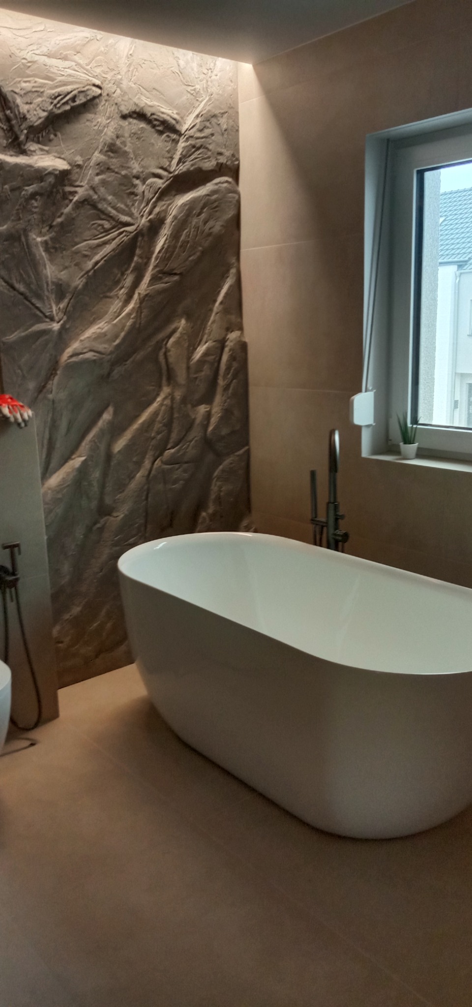 Nowoczesna łazienka z wolnostojącą wanną na tle ściany o fakturze imitującej skałę, oświetlona punktowo, z widokiem na budynek za oknem.