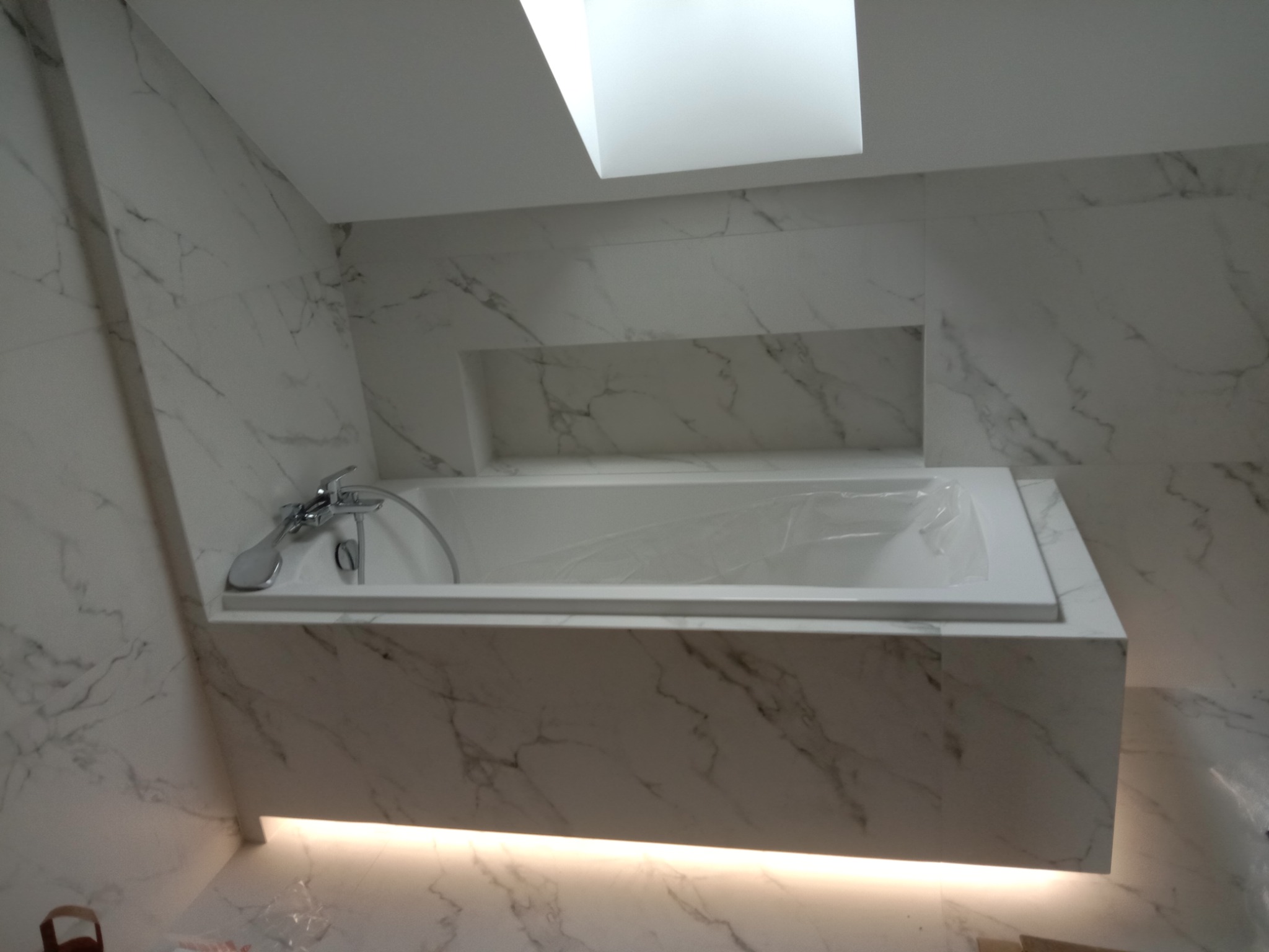 Nowoczesna łazienka z wanną obudowaną płytkami imitującymi marmur, podświetleniem LED u dołu i wnęką w ścianie. Minimalistyczny design, jasne kolory.