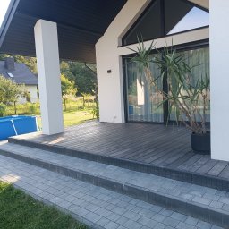 Strodrew - Nowoczesny taras drewniany z ciemnymi deskami, schody z kostki brukowej, białe filary i roślina w donicy. W tle dom z basenem.