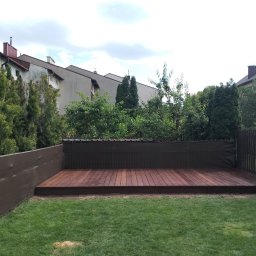 Strodrew - Wykończony taras z drewna egzotycznego w ogrodzie, otoczony zielenią i panelami osłonowymi. Widok na zabudowę miejską w tle, w dziennym oświetleniu.