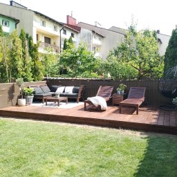 Strodrew - Przestronny taras z drewna w ogrodzie, z meblami wypoczynkowymi i leżakami. Widoczne budynki w tle oraz zieleń drzew i trawnik. Całość utrzymana w ciepłej tonacji.