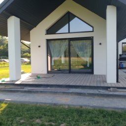 Strodrew - Nowoczesny dom z tarasem kompozytowym w trakcie budowy. Widoczne deski tarasowe, narzędzia i elementy konstrukcyjne. Jasna elewacja i duże przesuwne okna.