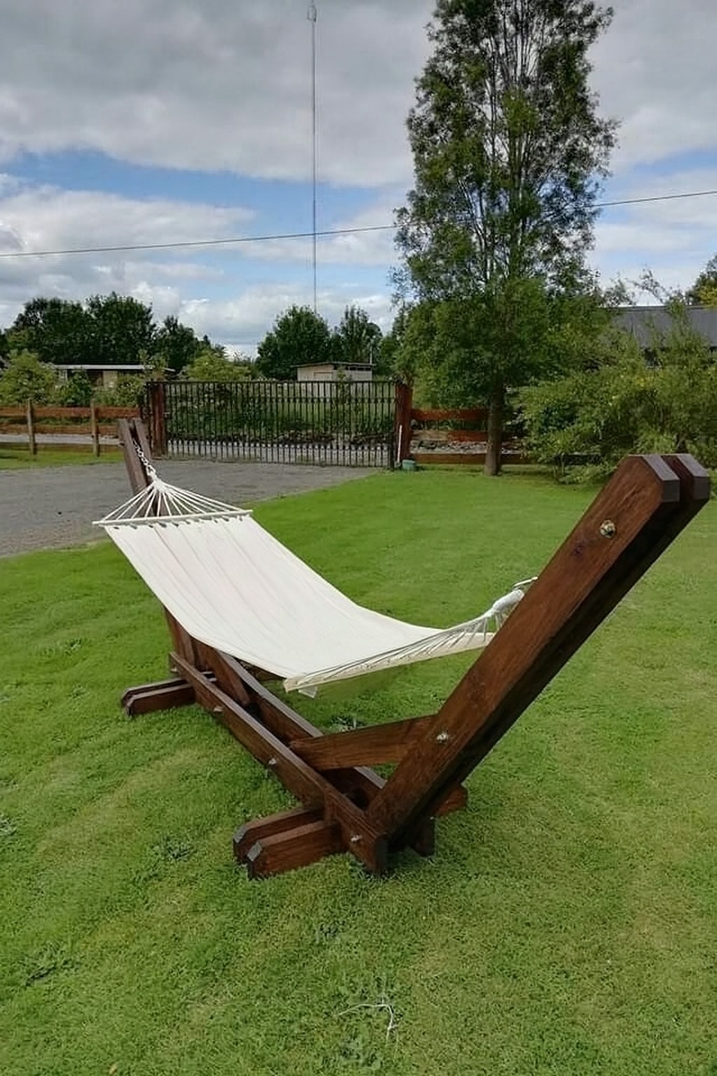Drewniany hamak z białym płótnem, umieszczony na trawniku przed domem. Widoczne detale konstrukcji z ciemnego drewna i metalowe śruby. W tle drzewa i ogrodzenie.