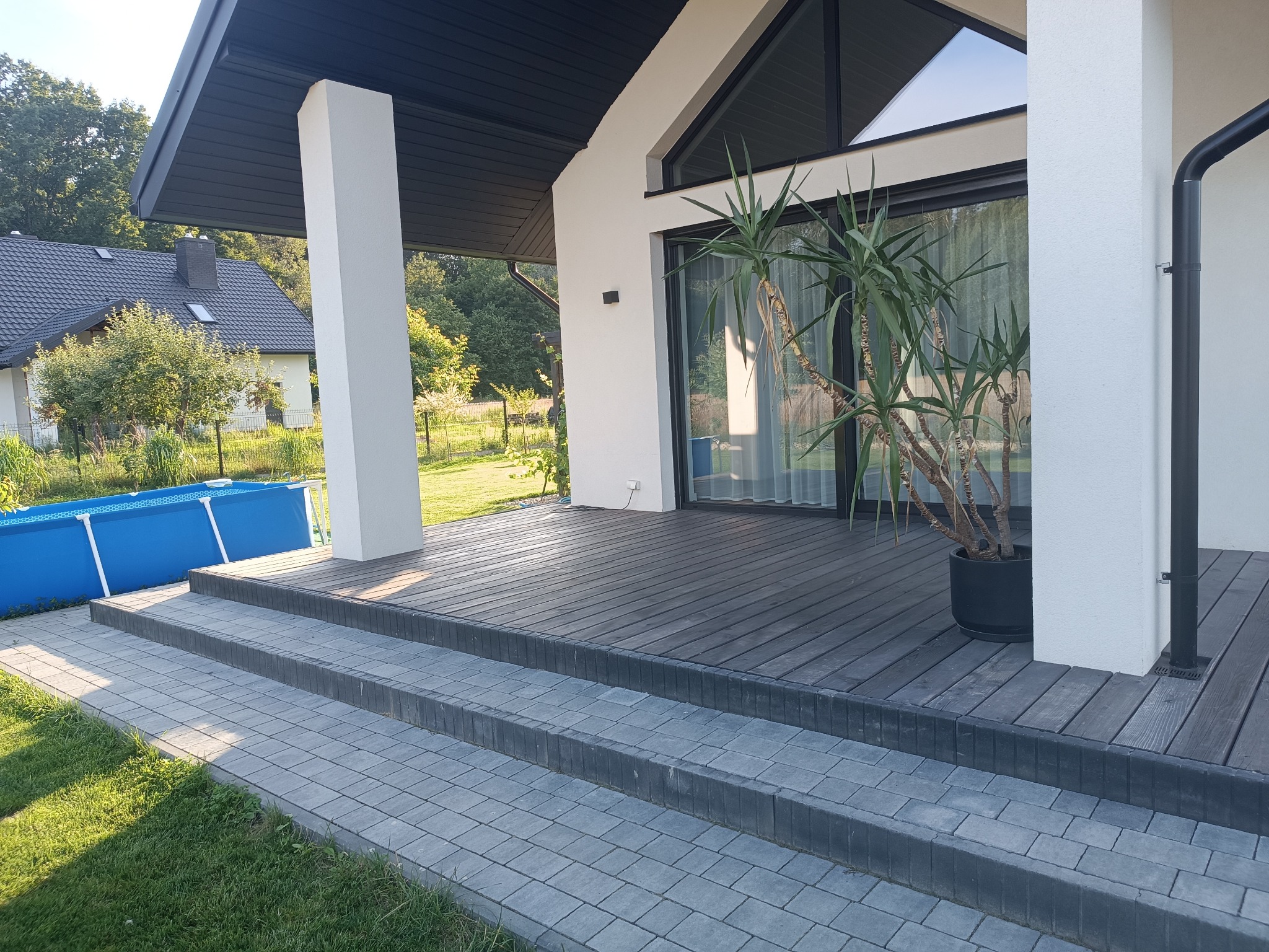 Nowoczesny taras drewniany z ciemnymi deskami, schody z kostki brukowej, białe filary i roślina w donicy. W tle dom z basenem.