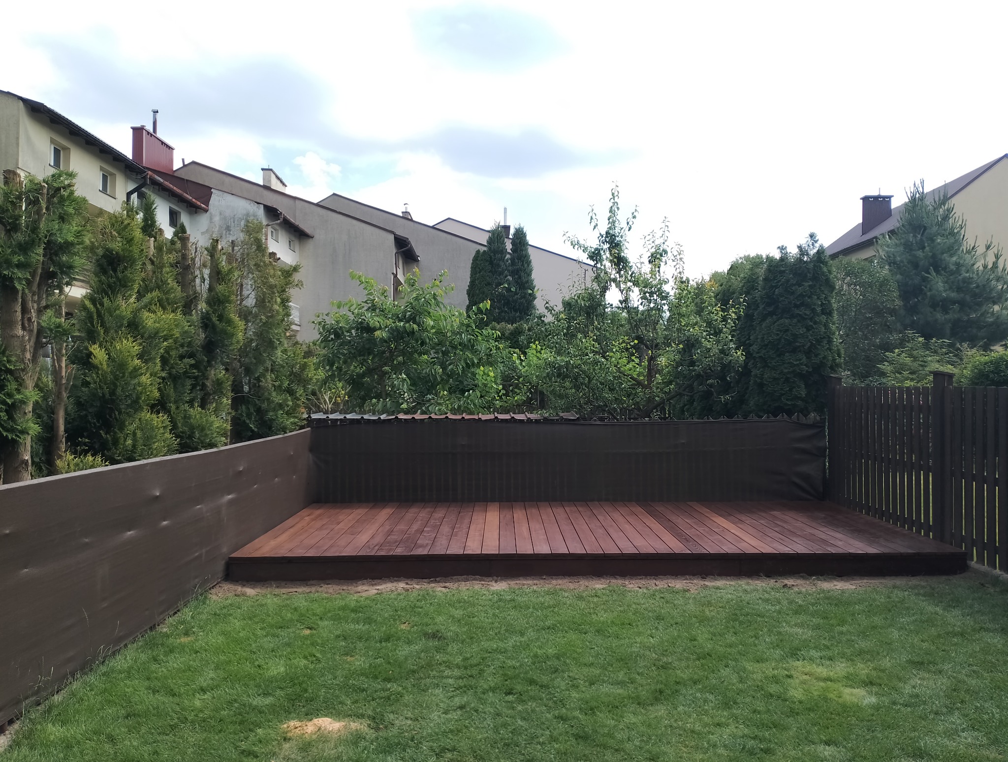 Wykończony taras z drewna egzotycznego w ogrodzie, otoczony zielenią i panelami osłonowymi. Widok na zabudowę miejską w tle, w dziennym oświetleniu.