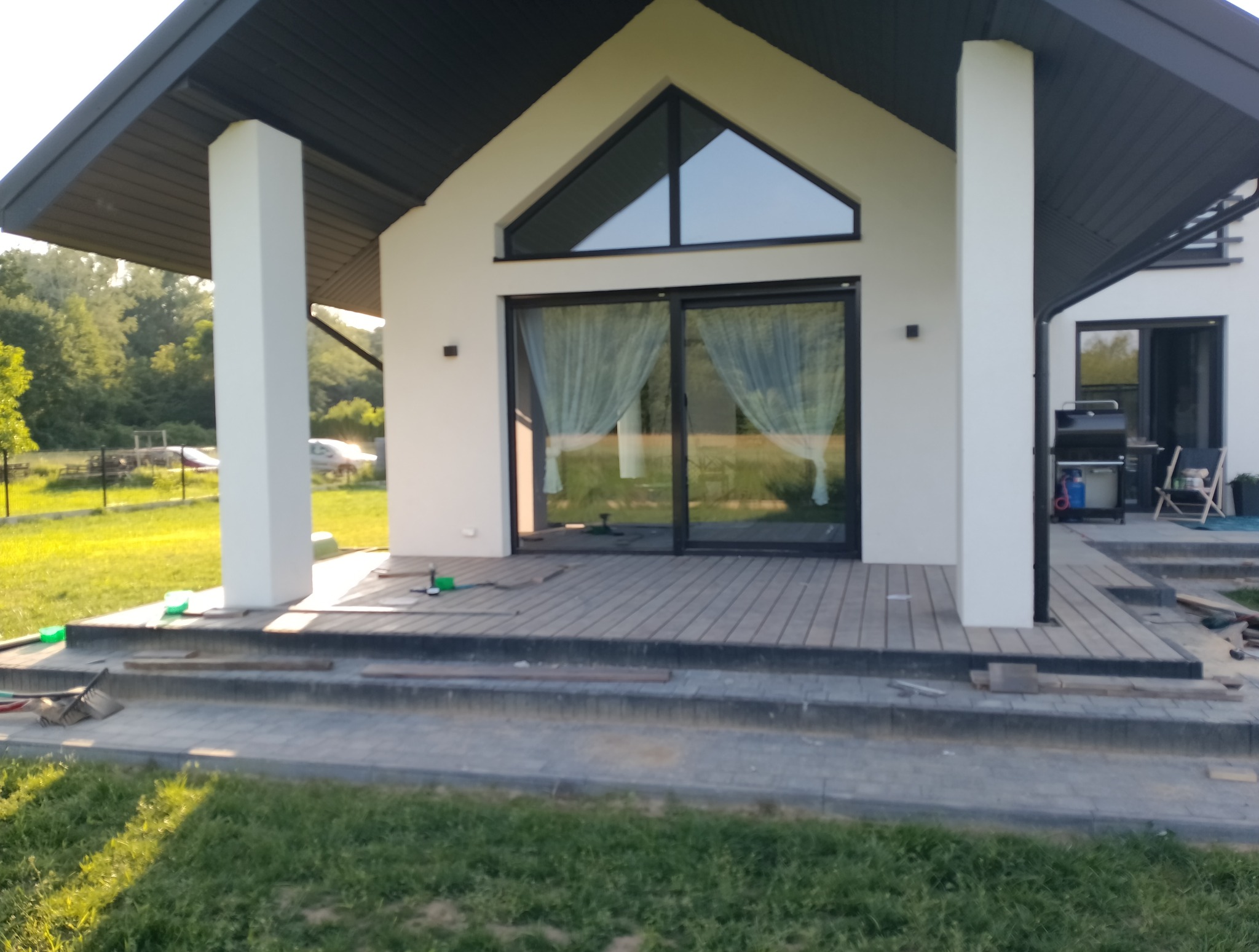 Nowoczesny dom z tarasem kompozytowym w trakcie budowy. Widoczne deski tarasowe, narzędzia i elementy konstrukcyjne. Jasna elewacja i duże przesuwne okna.