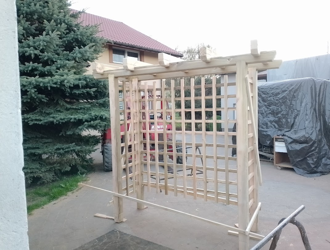 Surowa, drewniana konstrukcja kratownicy w ogrodzie, z widoczną maszyną budowlaną w tle. Etap budowy, jasne drewno, regularna siatka kraty, zielony świerk obok.