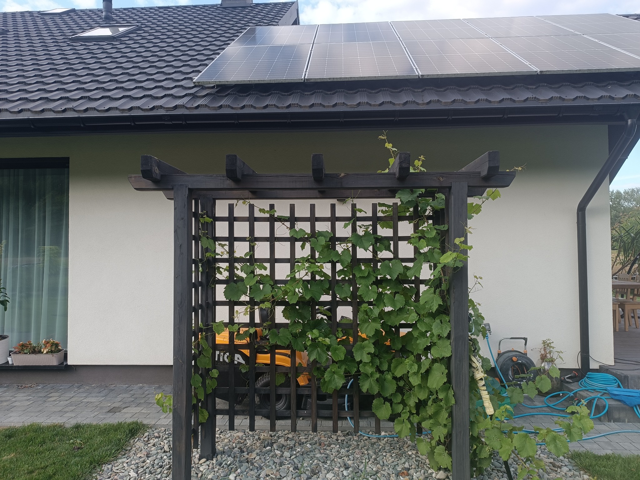 Ciemna, drewniana konstrukcja z kratownicą, obrośnięta zielonym winem, na tle jasnego domu z panelami słonecznymi. W tle widoczny żółty pojazd.