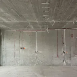 Surowy stan: Instalacja elektryczna w nowym budynku z betonowymi ścianami i widocznymi przewodami w kolorze białym i czerwonym. Przewody poprowadzone po suficie i ścianach.